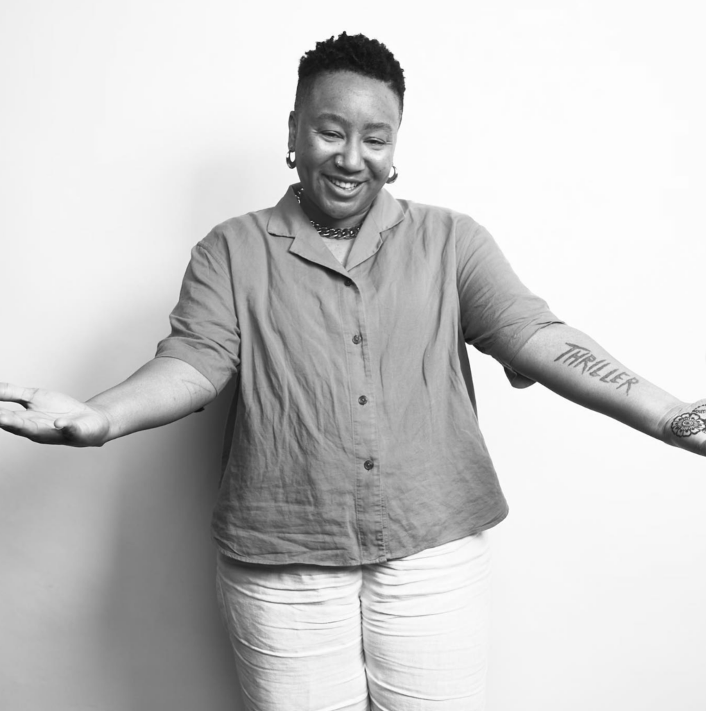 A black and white image of Stas THEE Boss against a white backdrop smiling with her arms outstretched towards the camera.