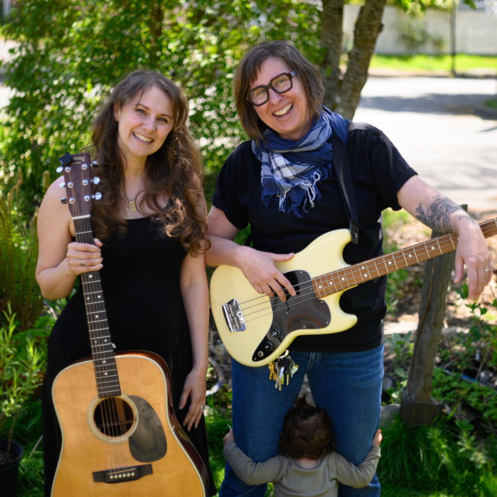 Artists Bannister & Maine, one holding an acoustic guitar on the left, and and the other holding an electric bass guitar on the right.