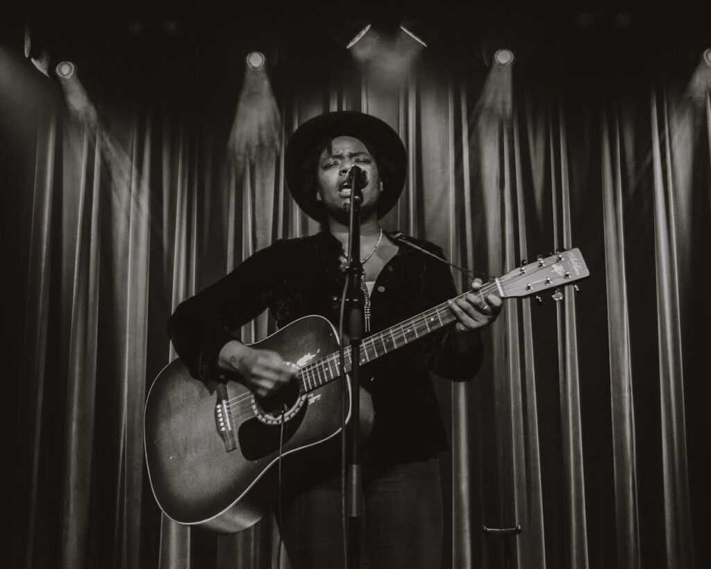 A sepia-toned photo of musician Maya Marie playing the guitar and singing towards the camera.