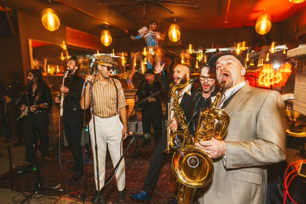 Members of the band Reposado in a red-tinted and dimly lit bar. One member in the center is standing holding a microphone and two other men on the right are playing saxophones.