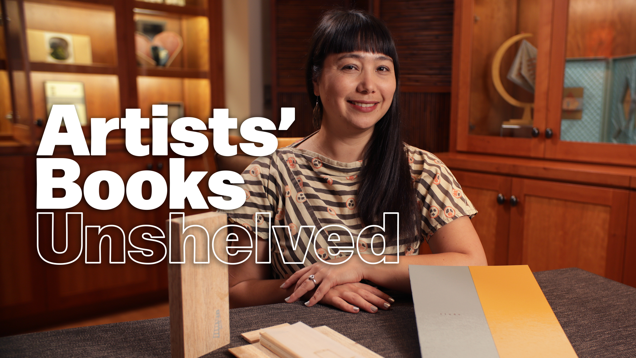 Artist and presenter Yuka Petz poses at a table with two artists' books in a warmly lit room with bookcases behind her