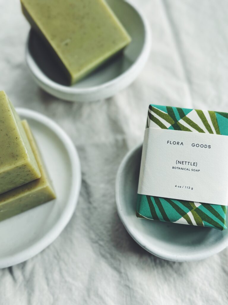 Green bars of soap sit on white plates on a white cloth, the label on one says Flora Goods, Nettle Botanical Soap