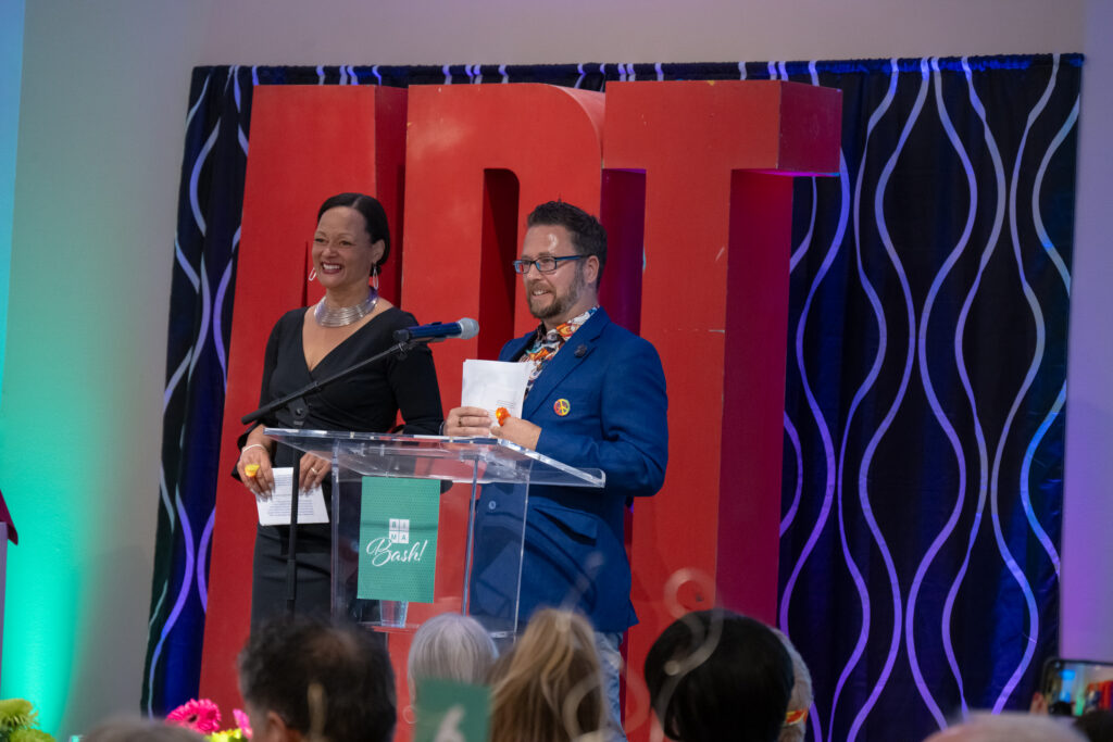 Two presenters stand at a clear podium on a stage, smiling as they address an audience with a colorful red backdrop behind them.