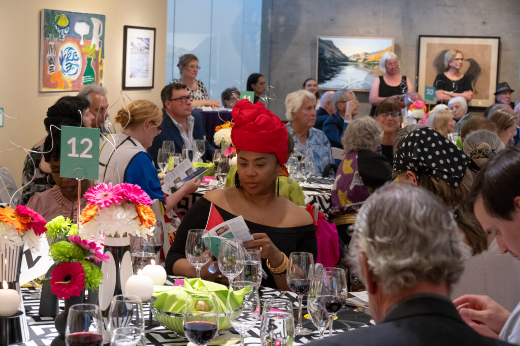 People seated at decorated round tables in an art gallery-style venue, guests mingle over wine as bright flowers and table numbers appear in the foreground.