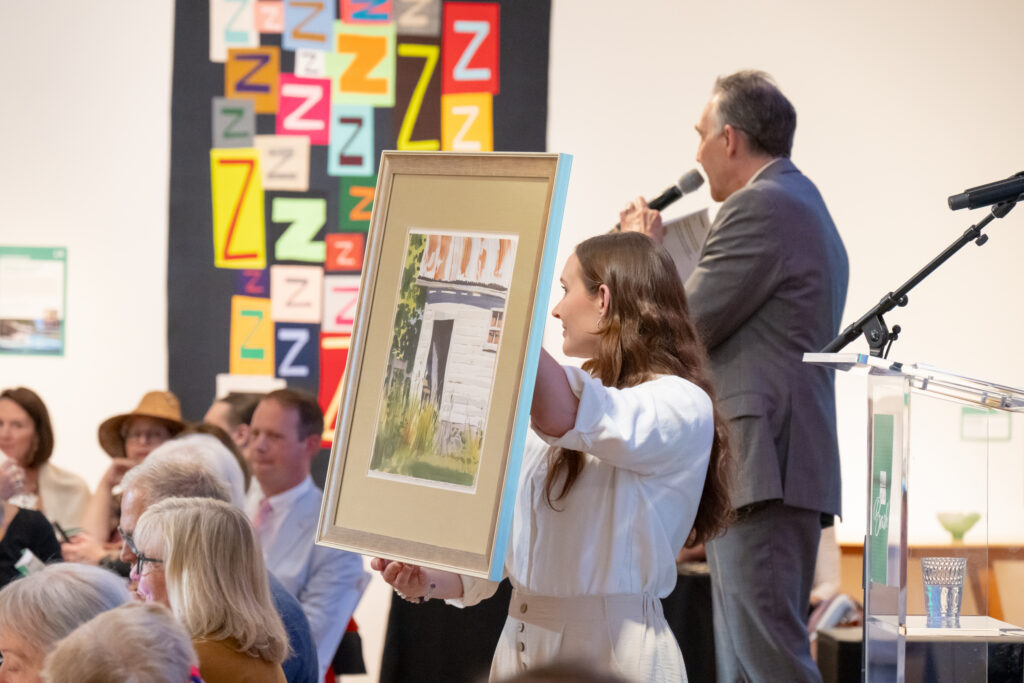 Woman in a white blouse holds a large framed painting bidily at an art auction, with a man speaking into a microphone behind her.
