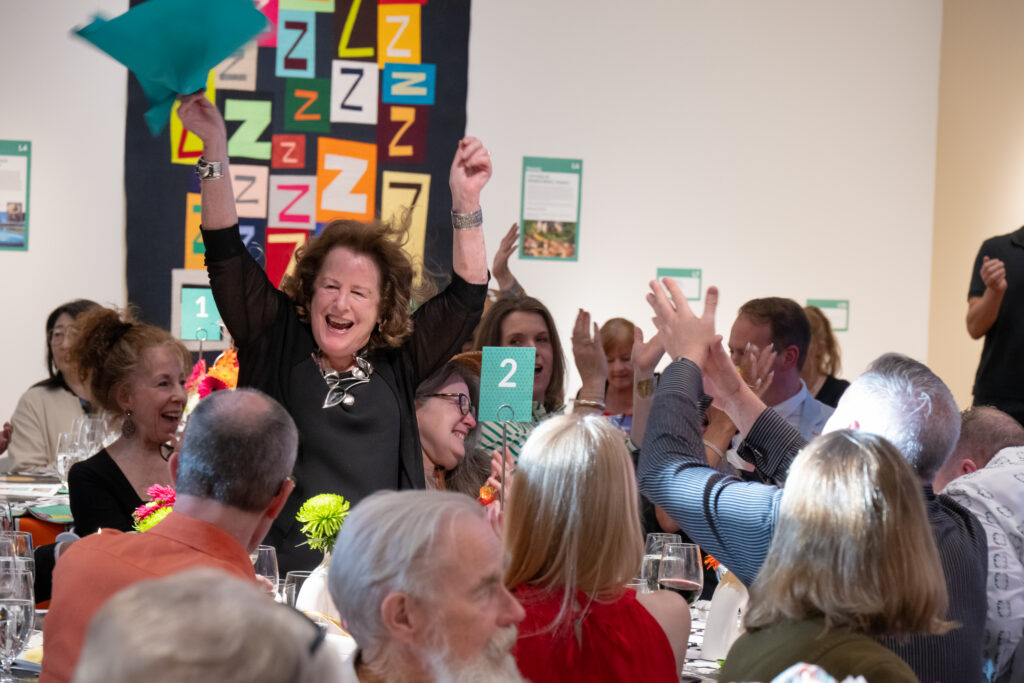 Celebration at a dinner event with a smiling woman raising arms while others cheer around tables with glasses and decorations behind her.