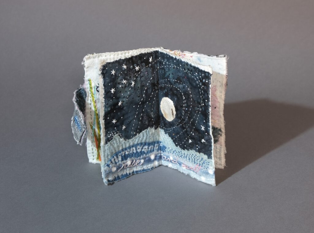 A small fabric book opened to show a dark stitched night sky with a full moon, surrounded by white embroidered stars and hand-stitched text at the bottom.