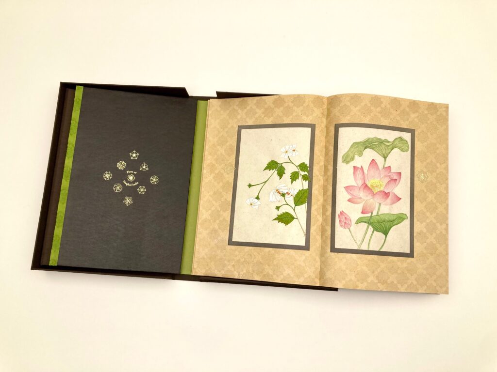 An open book in a black case with green trim. The two visible pages show botanical illustrations of white and pink flowers, each centered in a rectangular frame on a patterned tan background.