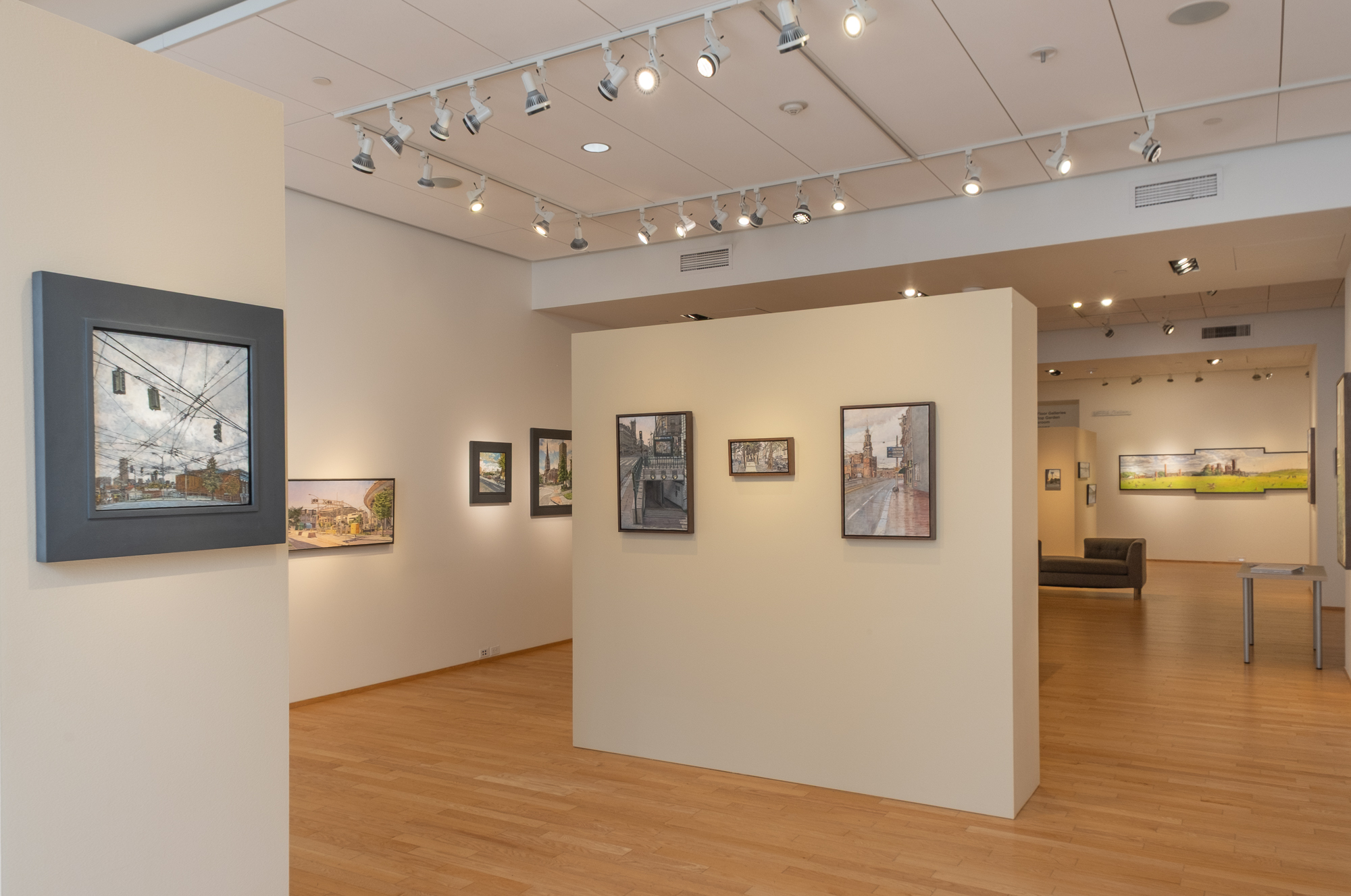 Gallery interior with framed urban landscape paintings on light-colored walls, wood floors, and ceiling track lighting; a gray couch and additional artwork are visible in the background.
