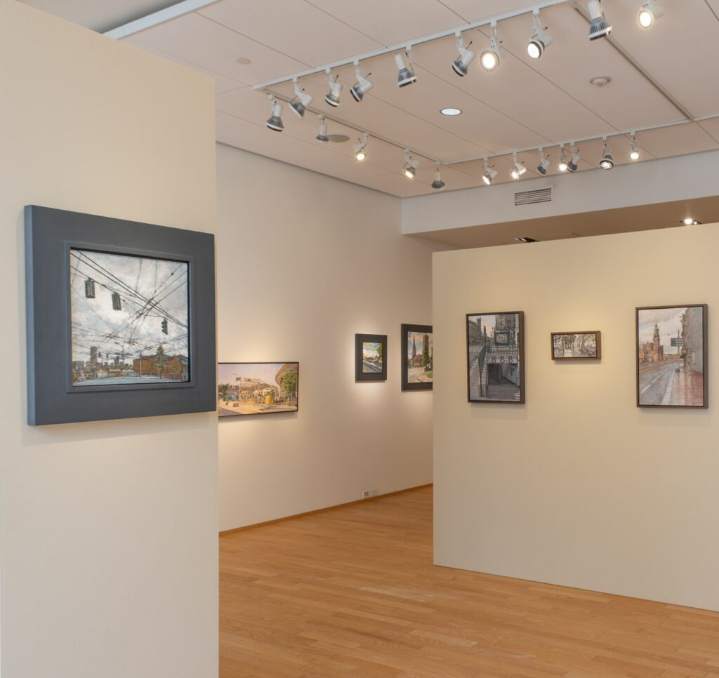 Gallery interior with framed urban landscape paintings on light-colored walls, wood floors, and ceiling track lighting; a gray couch and additional artwork are visible in the background.