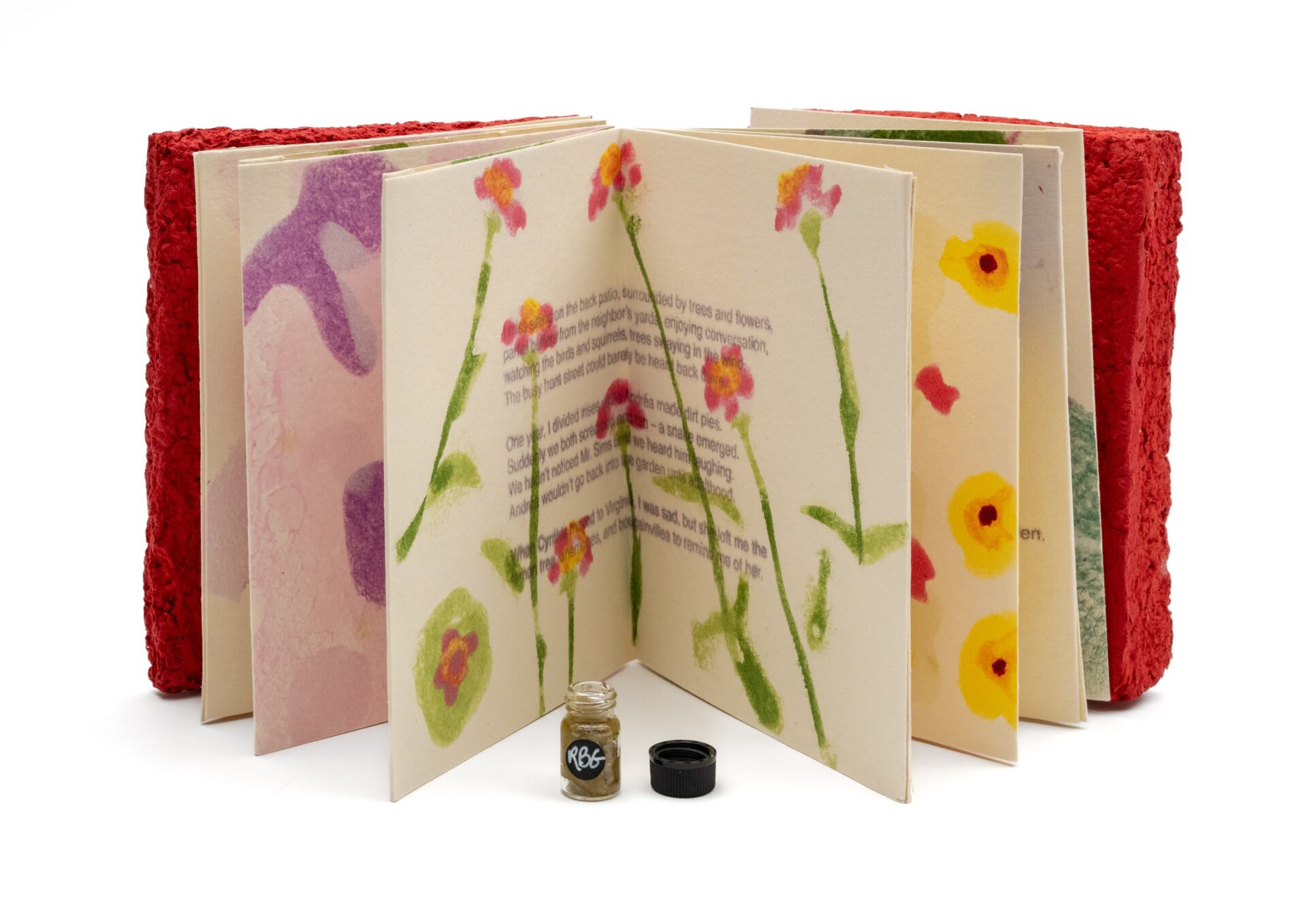 accordion-style book with brightly illustrated flowers, a thick red cover, sitting next to a vile filled with seeds