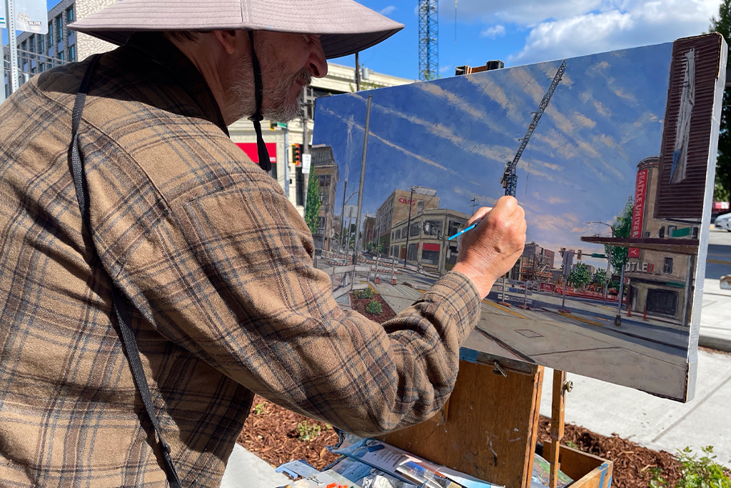 Man painting on a sidewalk wearing a bucket hat and plaid shirt