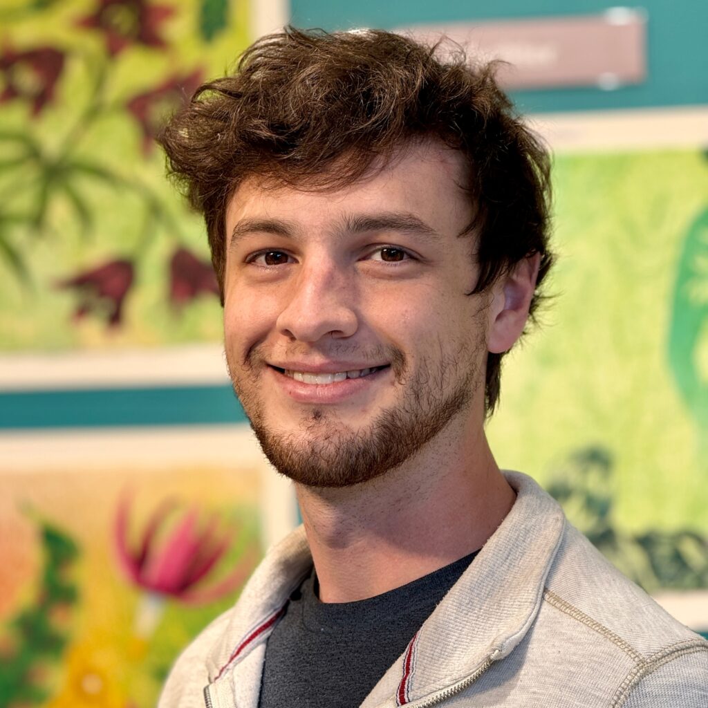 A young man with tousled brown hair and a trimmed beard smiles in front of a colorful wall of floral art. He is wearing a light hoodie over a dark T-shirt.