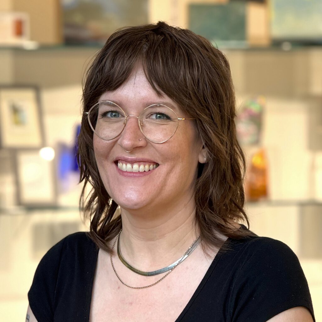 A woman with shaggy brown hair and glasses smiles in front of a softly lit display of artworks. She wears a black top and layered silver necklaces.