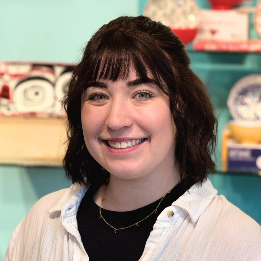 A young woman with shoulder-length dark brown hair and bangs smiles in front of a display of colorful ceramics. She is wearing a white button-up shirt over a black top, and layered gold necklaces.