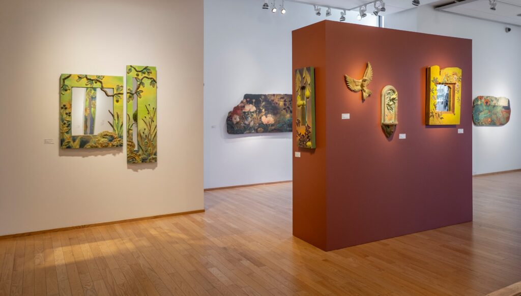 A wideshot of a museum gallery. On the left are mirrors in green, on the right is a red wall with concrete sculptures. In the background are floral paintings.