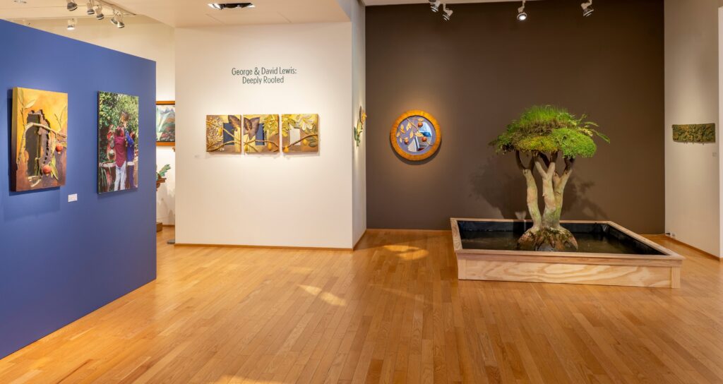 A wide shot of a museum gallery. On the left is a blue wall with garden photos. The back wall has mirrors and a concrete tree in a fountain.