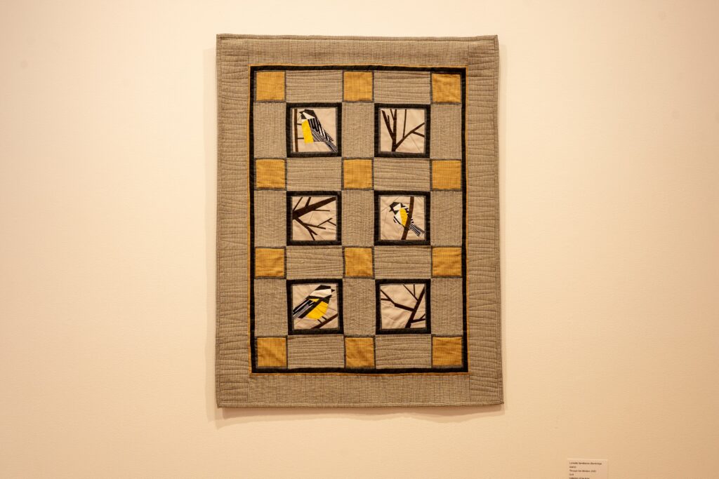 A grey quilt hangs on the wall with yellow squares and black white and yellow birds
