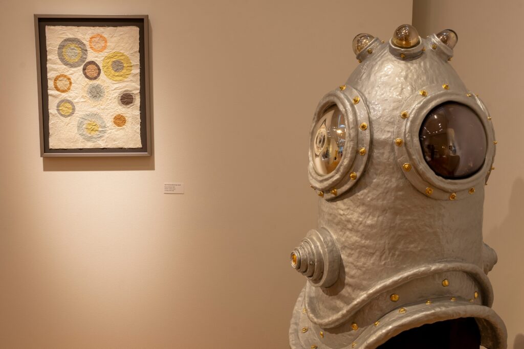 Foreground has a large ceramic silver diving helmet The background has a framed paper piece with circles