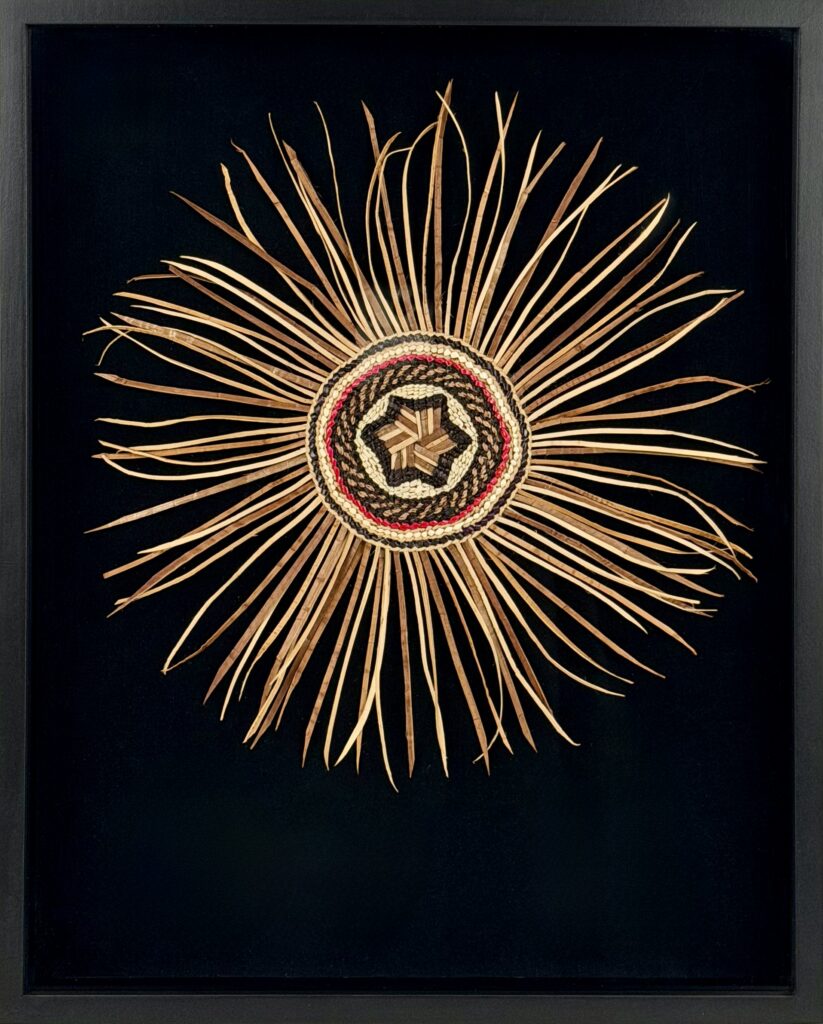 Decorative sunburst made from woven straw spokes radiating from a circular center on a black background in a frame