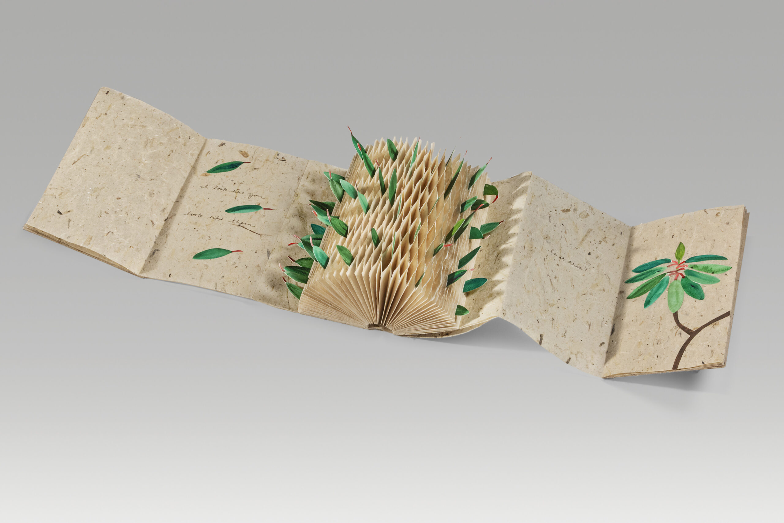 A tan, open accordion book that has green handmade leaves coming from the pages and on some of the pages.