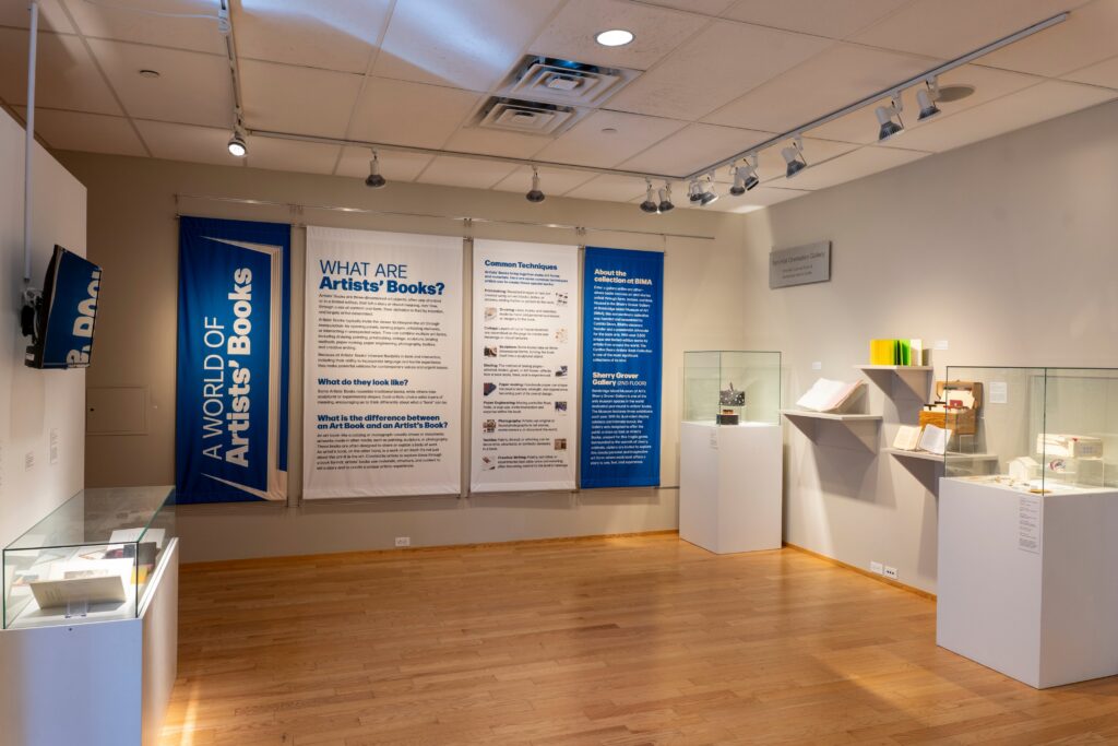 A wide shot of a museum gallery with artists books on white stands There are blue and white banners and one says a world of artists books 