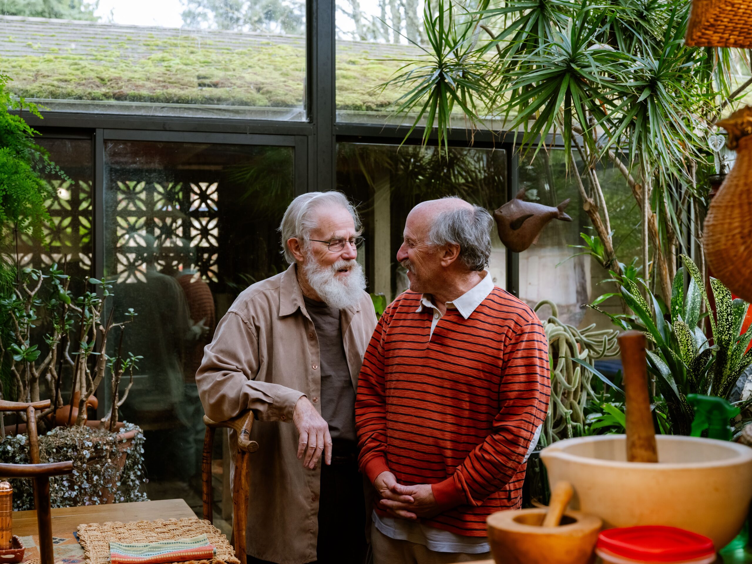Two white men stand in a green garden. One has a long white beard with a tan shirt and the other has an orange striped shirt on. They are looking at each other.