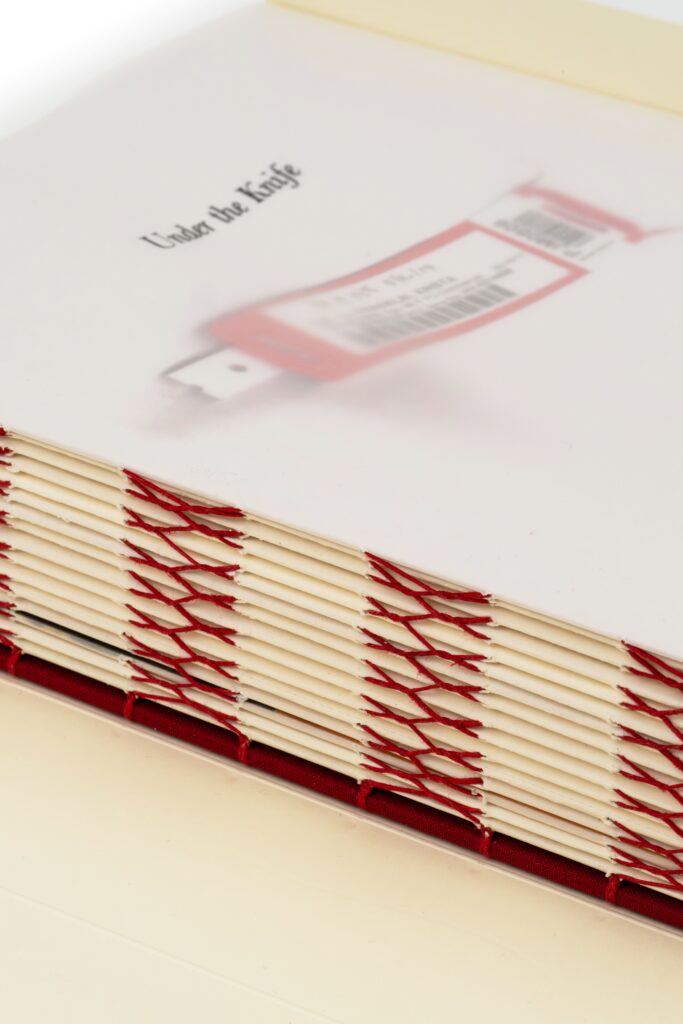 The binding of white pages threaded with red thread text on the top page says under the knife 