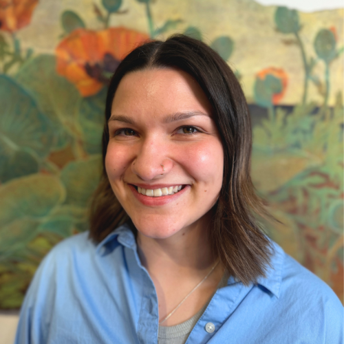Portrait of a smiling woman with shoulder length brown hair wearing a light blue button up shirt standing in front of a colorful mural