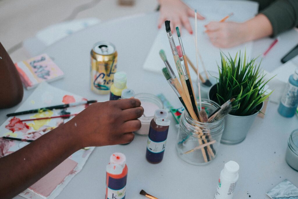 Child's hands painting at a table with brushes in a jar, paint tubes, and a plant nearby.