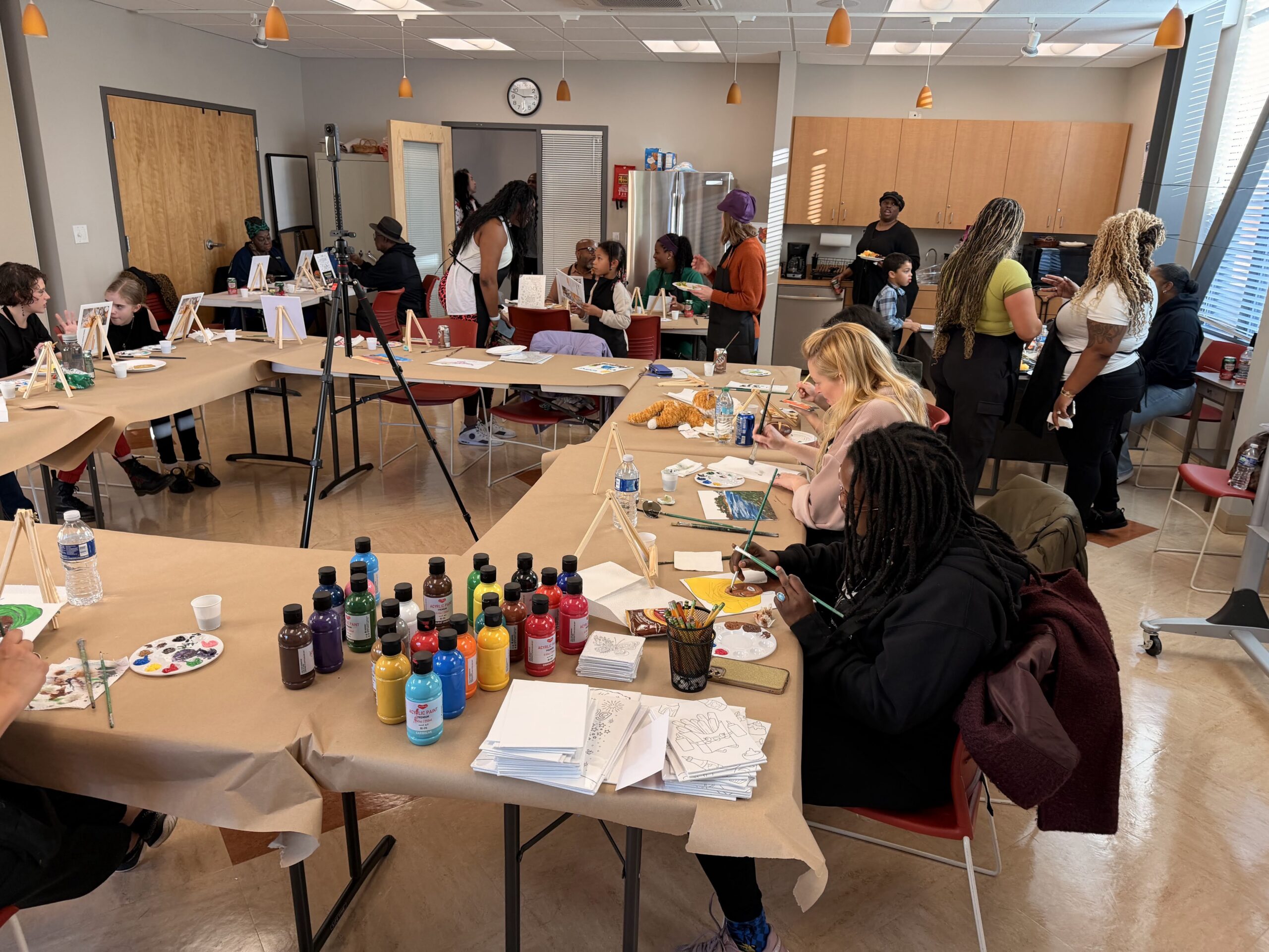People sit at long tables in a classroom painting, with many paint bottles on the tables.