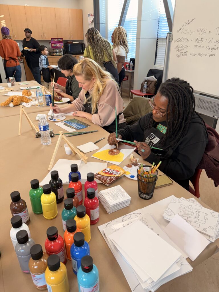 People sit at long tables in a classroom painting, with many paint bottles on the tables.